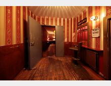 Ein schmaler Eingangsbereich mit Holzboden und vertikal gestreiften Tapeten in Rot- und Goldtönen. Eine geöffnete Metalltür führt in einen dahinterliegenden Raum mit gedämpfter Beleuchtung, links steht ein Tresen mit Hocker, und an der Wand hängen Schilder, darunter eines mit der Aufschrift "Kasse".