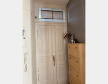 Weiße Holztür mit geometrischer Verzierung und einem Oberlicht mit eingefassten, dekorativen Milchglasscheiben. Rechts daneben steht eine helle Holzkommode mit darauf abgelegten Hüten und Utensilien; links an der Wand befindet sich eine Gegensprechanlage.