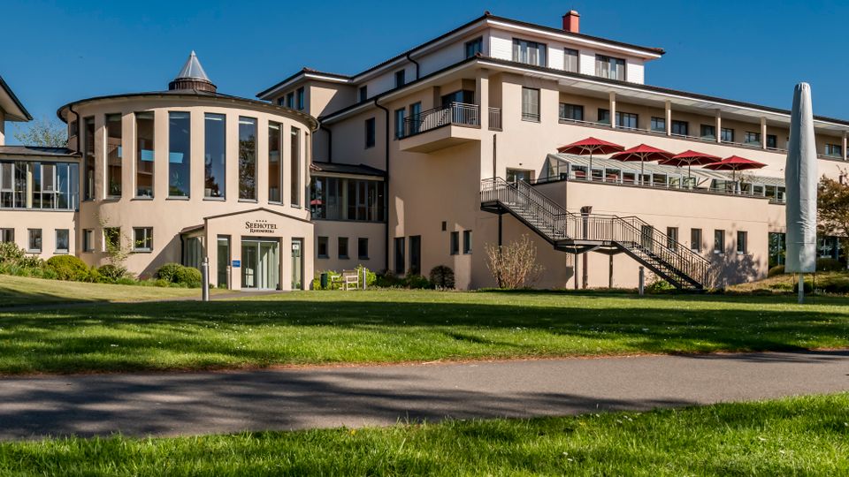 Exterior view of a modern multi-story hotel complex with a round glass-fronted central section, balconies, and an outdoor terrace with red umbrellas. The building sits beside a wide grassy lawn and a paved path under a clear blue sky.