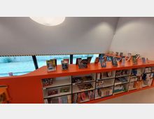 Ein moderner Innenraum einer Bibliothek mit einem orangefarbenen Regal, das im oberen Bereich Bücher auf Displayständern präsentiert. Im unteren Teil des Regals befinden sich Fächer mit weiteren Büchern, Comics und Spielen; im Hintergrund sind große Fenster und ein gepflasterter Außenbereich sichtbar.