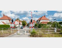 Bahnübergang in einer Wohngegend mit rot-weißen Schranken, Ampeln und Andreaskreuzen, umgeben von Einfamilienhäusern mit roten Ziegeldächern und gepflegtem Grün; Straßenschilder weisen auf die "Leibnitzstraße" und "Heinrich-Heine-Straße" hin. Im Hintergrund sind parkende Autos und Bäume zu sehen, unter einem bewölkten Himmel.