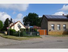 Straßenszene in einer ländlichen Gegend mit Einfamilienhäusern; rechts ein Haus mit Solarpanelen auf dem Dach, links ein helles Haus mit roten Ziegeldach. Dazwischen ein grüner Garten mit Zaun, Verkehrsschildern und dem Straßenschild „Dorfstraße“. Im Vordergrund gepflasterter Gehweg und Straße.