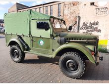 Grünes, militärisches Geländefahrzeug mit Plane, geparkt auf einem gepflasterten Platz vor einer alten, verfallenen Backsteinwand mit Graffiti. Die Umgebung wirkt urban und leicht verfallen.