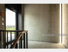 Treppenhausbereich mit Sichtbetonwänden, einer Metall- und Holzgeländerkonstruktion und seitlich integrierter LED-Lichtleiste. Links befindet sich eine Glastür, die nach außen zu einem begrünten Bereich führt.