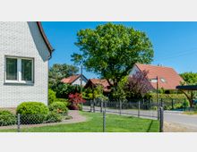 Wohnsiedlung mit Einfamilienhäusern, umgeben von gepflegten Gärten und großen Bäumen. Im Vordergrund ist ein weißes Haus mit einem Zaun und Rasenfläche, im Hintergrund weitere Häuser mit roten Ziegeldächern zu sehen.