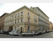 Ecke eines historischen Wohngebäudes mit hellgelber und grauer Fassade, zahlreichen symmetrisch angeordneten Fenstern mit Zierleisten. Auf der gepflasterten Straße im Vordergrund parken mehrere Autos.