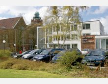 Außenansicht der Gottfried-Benn-Bibliothek in Berlin, einem modernen weißen Gebäude mit Glasfassade und roter Backsteinfront, umgeben von Bäumen und geparkten Autos. Im Hintergrund ist ein historischer Backsteinturm mit Kuppel zu sehen.