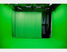 Ein Aufnahmestudio mit einem durchgehenden Greenscreen-Hintergrund, bestehend aus grün gestrichenen Wänden, einem grünem Boden und einem grünen Vorhang. Im oberen Bereich sind Studiobeleuchtung und Kabel sichtbar.