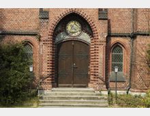 Eingang einer Backsteinkirche mit massiver Holztür, verziert mit geschmiedeten Metallelementen. Über der Tür ein rundes Mosaik mit einem Lammmotiv, flankiert von gotischen Fenstern mit Buntglas und Rankenmustern.