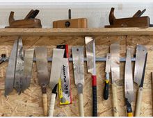 Wand mit einer Holzhalterung in einer Werkstatt, auf der verschiedene Handsägen und Hobel organisiert aufgehängt sind. Im Hintergrund ist eine Spanplatte als Wandverkleidung sichtbar.