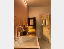 Kompakte Küche mit einer weißen Spüle im Vordergrund auf einer Holzarbeitsplatte, rosa Fliesenspiegel an der Wand, schmalem Regal mit Küchengeräten und einer geöffneten Holztür im Hintergrund. Eine kleine Tischlampe beleuchtet den Arbeitsbereich.