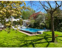 Grüne Gartenlandschaft mit einem großen rechteckigen Pool, umgeben von Bäumen und Pflanzen. Im Hintergrund ein weißes Wohnhaus mit roten Ziegeldächern und einer Terrasse, die mit Sitzmöbeln ausgestattet ist.