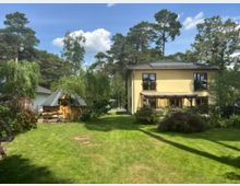 Großer, gepflegter Garten mit Rasenfläche, umgeben von Sträuchern und hohen Bäumen, mit einem zweistöckigen Wohnhaus mit Terrasse sowie einem kleinen Holz-Gartenhaus mit Schindeldach. Im Hintergrund sind weitere Häuser und ein blauer Himmel mit Wolken zu sehen.