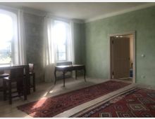 Ein heller Raum mit hellgrünen Wänden und zwei großen Fenstern mit weißen Vorhängen. Im Vordergrund befinden sich rote, gemusterte Teppiche, ein Schreibtisch mit geschwungenen Beinen sowie ein kleiner Tisch mit zwei Stühlen; im Hintergrund führt eine Tür zu einem angrenzenden Raum.