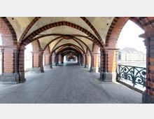 Blick durch einen überdachten Gang mit gotischen Bögen aus rotem Ziegelstein, Teil der Oberbaumbrücke in Berlin. Rechts ist ein Geländer mit floralem Muster sowie im Hintergrund ein modernes Gebäude zu sehen, links verlaufen Bahnschienen.