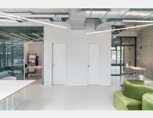 Moderner Büroraum mit minimalistischer Einrichtung, weiß gestrichenen Wänden und Glaselementen. Links ein Blick in ein separates Büro, rechts eine Sitzecke mit grünem Sofa, freiliegende Lüftungsrohre und lineare Deckenleuchten betonen den industriellen Stil.