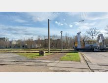 Industrielle Außenanlage mit Gleisen, einem gelben Schienenfahrzeug und Betonwegen. Im Hintergrund stehen Bäume und verfallene Gebäude, darunter eine graue Halle mit Fenstern.