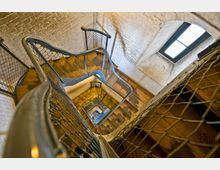 Schmale Wendeltreppe mit Holzstufen und Metallgeländer, umgeben von weiß gestrichenen Ziegelwänden. Rechts oben ein rechteckiges Fenster mit schlichtem schwarzem Rahmen, das natürliches Licht einfallen lässt.