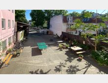 Ein gepflasterter Innenhof mit Tischtennisplatte, Holzbänken, Fahrrädern und mehreren Pflanzentöpfen. Im Hintergrund sind Wohnhäuser, ein kleiner Lagerbereich und Bäume sichtbar.