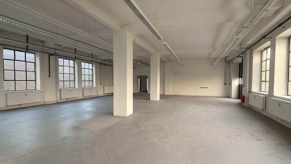 Großer, leerer Industrie-Loft-Raum mit Betonboden, weißen Stützsäulen und hohen Decken mit sichtbaren Kabeltrassen und langen Leuchten. Entlang der Wände befinden sich große, rasterförmige Fenster und Heizkörper; im Hintergrund ist eine Türöffnung zu sehen.