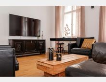 Wohnzimmer mit modernem Interieur, bestehend aus einem großen Flachbildfernseher an der Wand, einem verzierten dunklen Holzschrank darunter, schwarzen Ledersesseln, einem Holzcouchtisch und einem Schreibtisch vor einem Fenster mit beigen Vorhängen. Im Raum befinden sich dekorative Elemente wie kleine Laternen auf dem Tisch und ein Kissen mit goldfarbenem Muster.