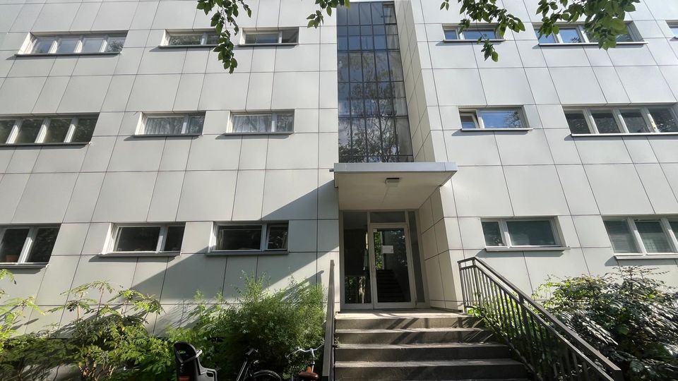 Eingangsbereich eines modernen, mehrstöckigen Gebäudes mit hellgrauer Fassadenverkleidung und Reihen von rechteckigen Fenstern. Eine Treppe mit Geländer führt zu einer verglasten Tür unter einem kleinen Vordach, umgeben von Büschen und überhängenden Baumzweigen.