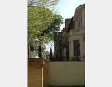 Eine Szene mit zwei Gebäuden: Im Vordergrund ein gelbes Backsteingeländer mit weißen Laternen und einer beigen Mauer. Im Hintergrund ein helles Gebäude mit klassizistischer Architektur und Balkonelementen sowie ein älteres, dunkleres Gebäude hinter Bäumen und Hecken.