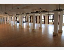 Großer leerer Raum mit Holzboden, weißen Säulen, bodentiefen Fenstern und weinroten Vorhängen. Die Raumdecke ist hoch, und an der Oberseite der Säulen sind Beleuchtungsschienen angebracht.