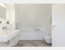 Helles, modernes Badezimmer mit schräger Dachdecke, eingebauter Badewanne in weißer Fliesenverkleidung und großformatigen grauen Bodenfliesen. Links ein breites Waschbecken mit Spiegel und Handtuchhalter, rechts ein wandhängendes WC neben einer offenen Tür.