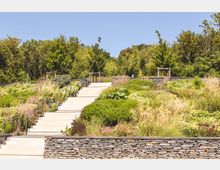 Steintreppe inmitten eines terrassierten Gartens mit üppiger Bepflanzung aus Gräsern, Sträuchern und Blumen, umgeben von Bäumen; im Hintergrund hölzerne Pergolen und blauer Himmel.