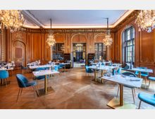 Elegantes Restaurant in einem historischen Raum mit holzgetäfelten Wänden und goldenen Verzierungen. Kristallleuchter hängen von der hohen Decke, Marmortische und blaue Polsterstühle stehen auf einem Fischgrätparkettboden.