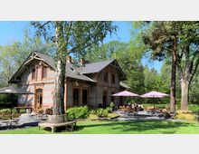 Backsteingebäude im Landhausstil mit grauem Dach, umgeben von grünen Bäumen und einer Rasenfläche. Im Vordergrund befindet sich ein Außenbereich mit Holztischen, Stühlen und Sonnenschirmen, teils besetzt von Gästen.