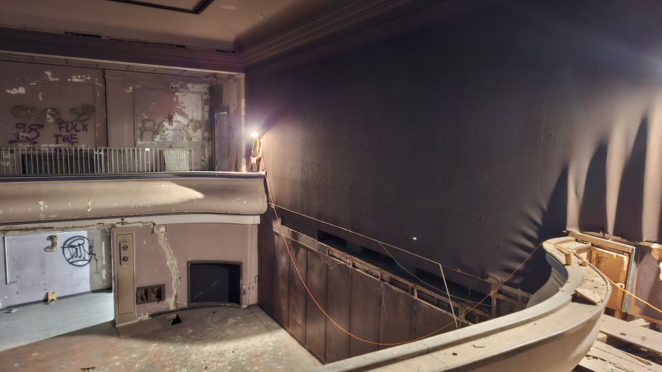 Verlassener Theater- oder Kinosaal mit leerer Bühne, einem Balkon mit Geländer und stark abgenutzten, teilweise abgeplatzten Wänden. Rechts dominiert eine große dunkle Wand mit freiliegenden Kabeln und Baustellenleuchte; der Boden ist staubig und stellenweise beschädigt.