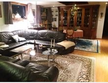 Wohnzimmer mit gemütlichem Ambiente, ausgestattet mit einer großen schwarzen Ledercouch in U-Form und einem Glastisch in der Mitte. Im Hintergrund befinden sich ein großer Holzschrank mit Glastüren, ein Esstisch mit einem runden Tischtuch und Teppiche auf hellem Holzboden.