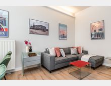 Modernes Wohnzimmer mit grauem Ecksofa und farbigen Kissen, kleinem Glastisch mit roter Ablage sowie hellem Holzboden. An den weißen Wänden hängen mehrere gerahmte Poster; daneben stehen ein schmaler Heizkörper und ein kleiner Beistelltisch mit Vase.