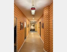 Flur mit Retro-Einrichtung, geprägt durch orange-gemustertes Tapetendesign und runde Wandecken. An der Decke hängen mehrfarbige, vintage-inspirierte Leuchten, und am Ende des Flurs ist ein Raum mit Möbeln im Retro-Stil sichtbar.