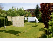 Eine grüne Rasenfläche in einem Garten mit Wäscheleinen, an denen bunte Bettlaken und Tücher hängen, umgeben von Bäumen, Sträuchern und einem roten Gebäudedach im Hintergrund.