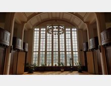 Großer Raum mit hohen, bunten Glasfenstern, die den Großteil der Rückwand einnehmen. Der Raum hat eine gewölbte Decke mit dekorativen Elementen, hölzerne Wandverkleidungen und eingebaute Emporen. Pflanzen und Möbel befinden sich entlang der Fensterfront.