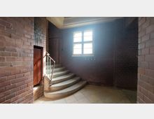 Treppenhaus mit bogenförmiger Steintreppe, umgeben von rotbraunem Mauerwerk. Ein großes Fenster mit geteilten Scheiben lässt Tageslicht in den Raum, rechts daneben befinden sich Haken an der Wand.