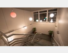 Treppenhaus mit modernem Design, beleuchtet durch kugelförmige, hängende Lampen in verschiedenen Farben. Große Fenster lassen nachts schwaches Außenlicht herein, und Zimmerpflanzen stehen dekorativ an den Wänden.