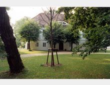 Historisches Gebäude mit weißen Wänden und einem mit roten Ziegeln gedeckten Dach, umgeben von gepflegtem Rasen und Bäumen. Ein kleiner, gestützter Baum steht im Vordergrund, und ein Kiesweg führt zum Eingang des Hauses.