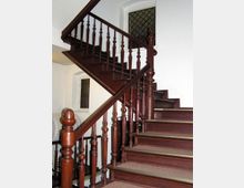 Holztreppe mit dunkelbraunem Geländer und gedrechselten Balustern in einem Innenraum mit weiß gestrichenen Wänden; im Hintergrund ein Fenster mit einem Gitter.