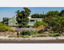 Flaches, modernes Gebäude mit grüner Fassade und farbigen Fensterläden, umgeben von üppiger Vegetation und einem angelegten Garten mit Kies, Sträuchern und Gräsern im Vordergrund; im Hintergrund hohe Bäume unter blauem Himmel.