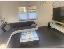 Modernes Wohnzimmer mit großem L-förmigem grauen Ecksofa, Glas-Couchtisch auf rundem Teppich und dunklem Fliesenboden. An der Wand befinden sich ein Fenster mit Plissees sowie ein wandmontierter Fernseher über einem weißen Lowboard vor einer grauen Steinoptik-Akzentwand.