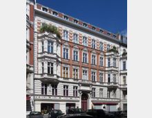 Fassade eines historischen Wohngebäudes im Stil des Spätklassizismus mit roten Backsteinwänden, weißen Putzverzierungen und Erkern. Das Gebäude hat mehrere Etagen, einen dekorativen Eingang mit rundem Torbogen und Dachfenster unter einem geneigten Dach.