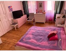 Ein gemütliches Schlafzimmer mit Holzdielenboden, einem Bett mit rosa und violetter Bettdecke, mehreren Kissen, und weißen Möbeln, darunter Kommoden und ein Schreibtischstuhl. Der Raum hat pastellrosa Vorhänge vor großen Fenstern, die von schwarzen Vorhängen eingerahmt werden.