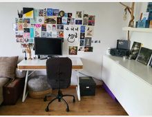 Ein Arbeitszimmer mit einem Schreibtisch, einem schwarzen Drehstuhl und einem Computerbildschirm. An der Wand hinter dem Schreibtisch ist eine Collage aus Postkarten und Bildern angebracht. Rechts steht ein weißes Sideboard mit Büchern, einer Musikanlage und Dekoration.