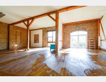 Großer, heller Raum mit einer Kombination aus freiliegenden Ziegelwänden, Holzbalken und einem glänzenden Holzboden. Große Fenster und eine Glastür im Hintergrund lassen viel Tageslicht herein; der Raum ist minimal möbliert mit einem blauen Sessel und einer Leiterregal-Dekoration.