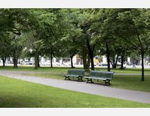 Ein Stadtpark mit grünen Bäumen, Rasenflächen und gepflasterten Wegen. Im Vordergrund stehen grüne Parkbänke, im Hintergrund sind Geschäfte, Autos und Menschen auf einer belebten Straße sichtbar.