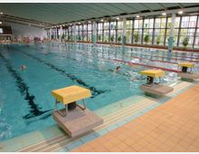 Hallenbad mit einem großen Schwimmbecken, das in mehrere Bahnen unterteilt ist, sowie Startblöcken am Beckenrand. Die Wände bestehen größtenteils aus Fenstern, durch die Tageslicht hereinfällt, und im Hintergrund sind Schwimmer und Pflanzen sichtbar.